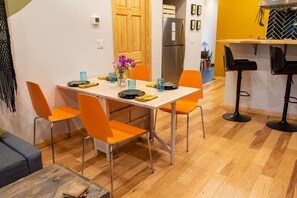 Comedor con una mesa preparada para cuatro personas, sillas naranjas alrededor, zona de cocina con refrigerador y barra con taburetes.