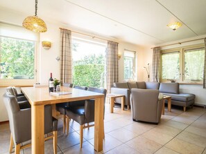 A modern living room with a dining area, featuring a sofa, chairs, a wooden table, and large windows with curtains. - Pet-free Texel Villa With Sauna (De Koog)