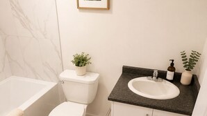 A modern bathroom with a toilet, sink, and bathtub. There is a framed picture on the wall and a small plant on the sink counter. - Home away from home (Edmonton)