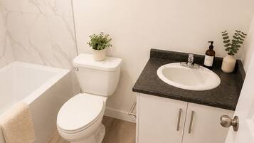 A modern bathroom with a toilet, sink, and bathtub. There is a framed picture on the wall and a small plant on the sink counter.