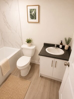 A modern bathroom with a toilet, sink, and bathtub. There is a framed picture on the wall and a small plant on the sink counter. - Home away from home (Edmonton)