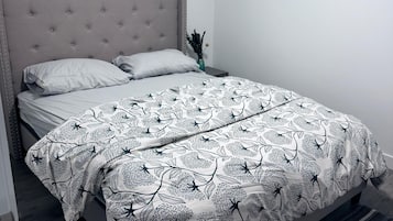 A bedroom with a bed, a window with blinds, a framed picture on the wall, and a bedside table with a vase.
