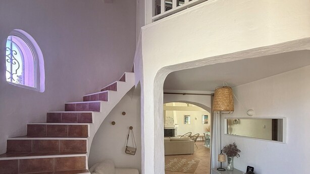 Een trap die leidt naar een tweede verdieping, met een witte balustrade, een klein raam met decoratieve ijzerwerk en een console tafel met een spiegel en decoratieve objecten.