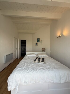 Un dormitorio con una cama grande, paredes blancas, suelo de madera, techo inclinado y una puerta que da a otra habitación.