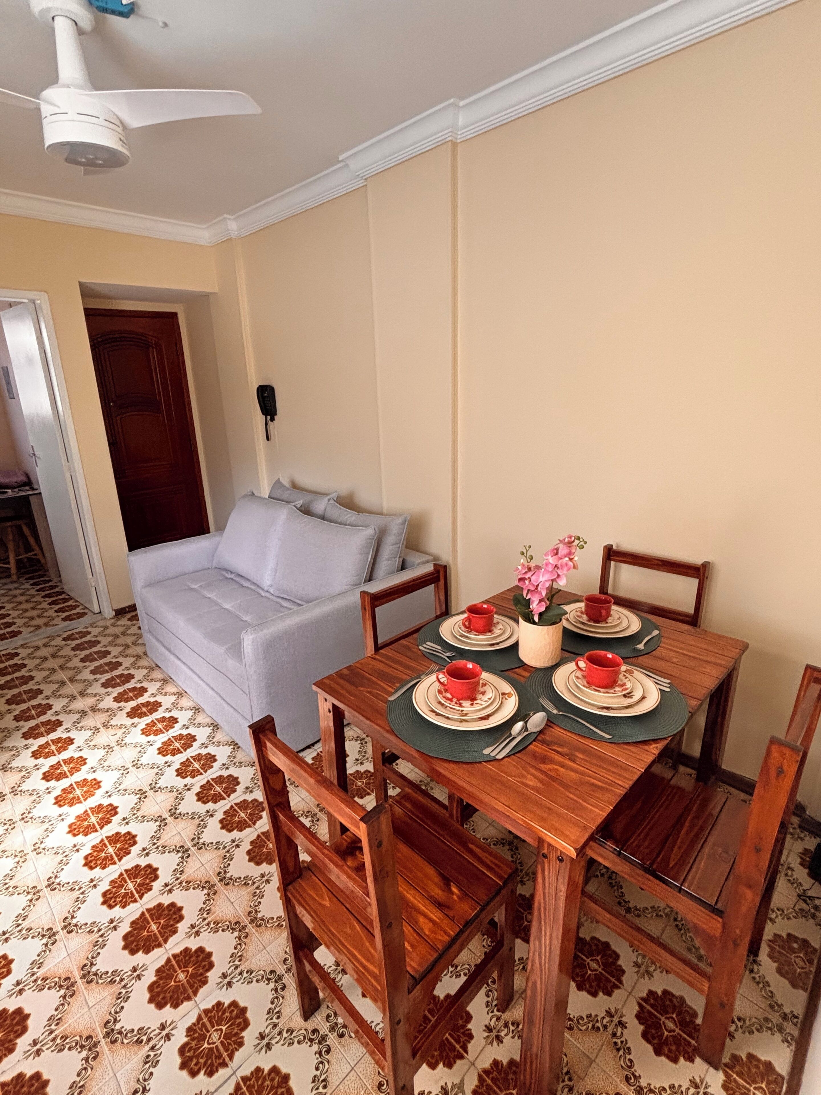 Un comedor con una mesa de madera preparada para cuatro personas, un sofá gris y un ventilador de techo.
