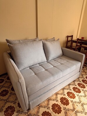 A grey sofa with a cushion, positioned against a wall with a patterned floor.