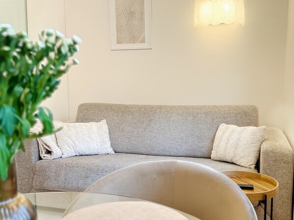 A neutral-toned sofa with two pillows, a small wooden side table, and a vase with green foliage.