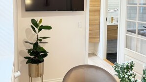 A modern living room with a flat-screen TV, a potted plant, a glass table, and a view of a kitchen area. - Det Gamle Vaskeri - Historical Apartment in Horsens (Horsens)
