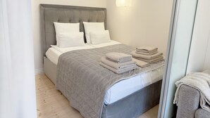 A neatly made bed with a grey headboard and white bedding, a stack of folded towels, and a light-colored wall with a single light fixture. - Det Gamle Vaskeri - Historical Apartment in Horsens (Horsens)