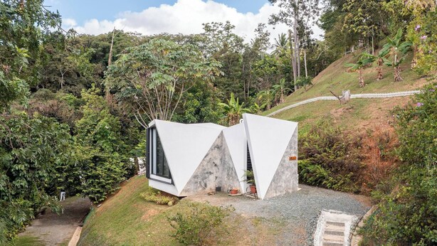 A modern house with a unique triangular roof design, surrounded by lush greenery and a white fence.