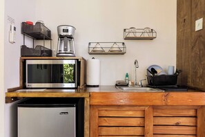 Cocina compacta con microondas, cafetera y refrigerador. Hay estantes con vajilla y un lavamanos pequeño con utensilios.