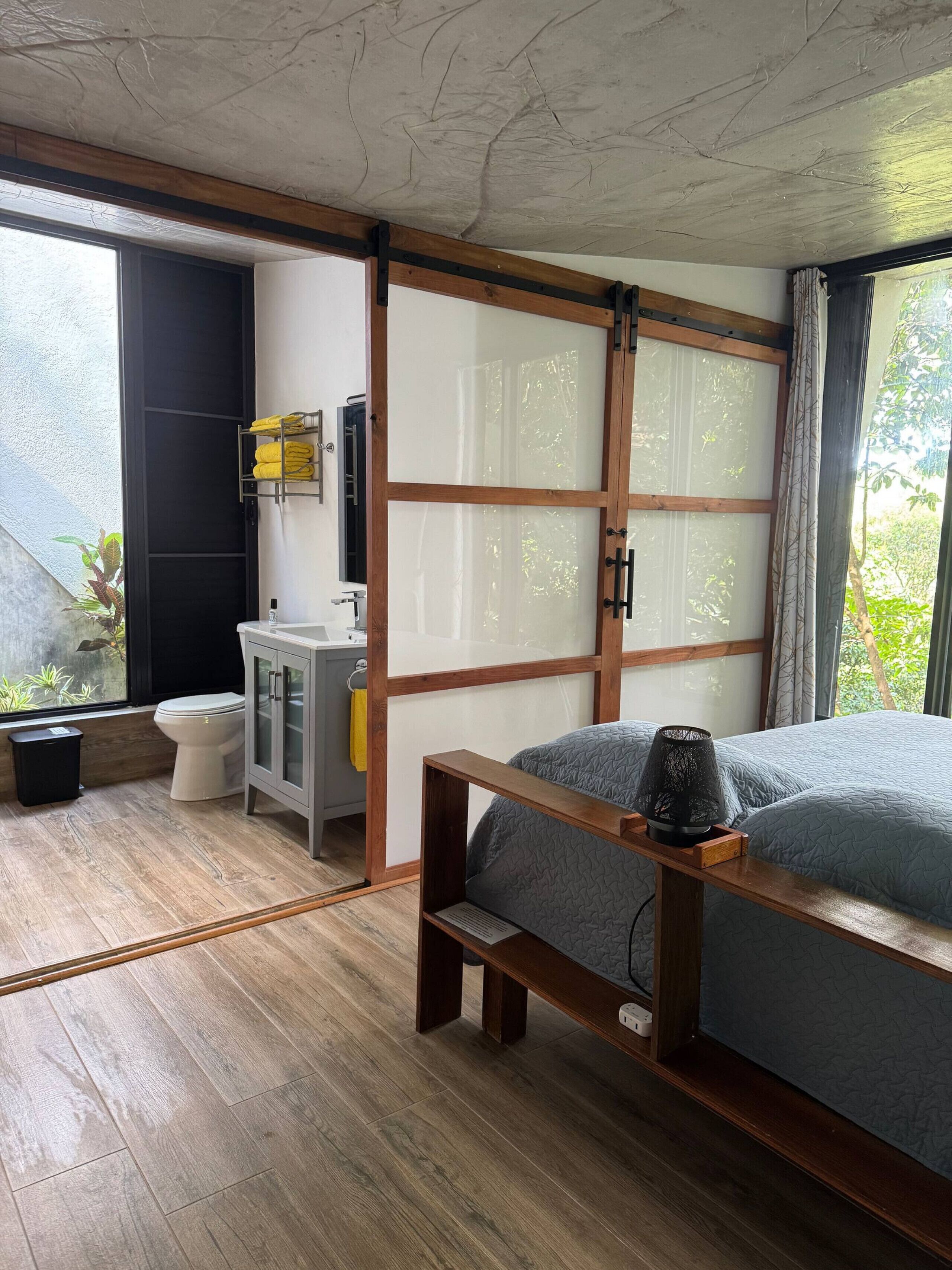 Ein Schlafzimmer mit Bett, Nachttisch und einem Fenster mit Blick auf Grünfläche.