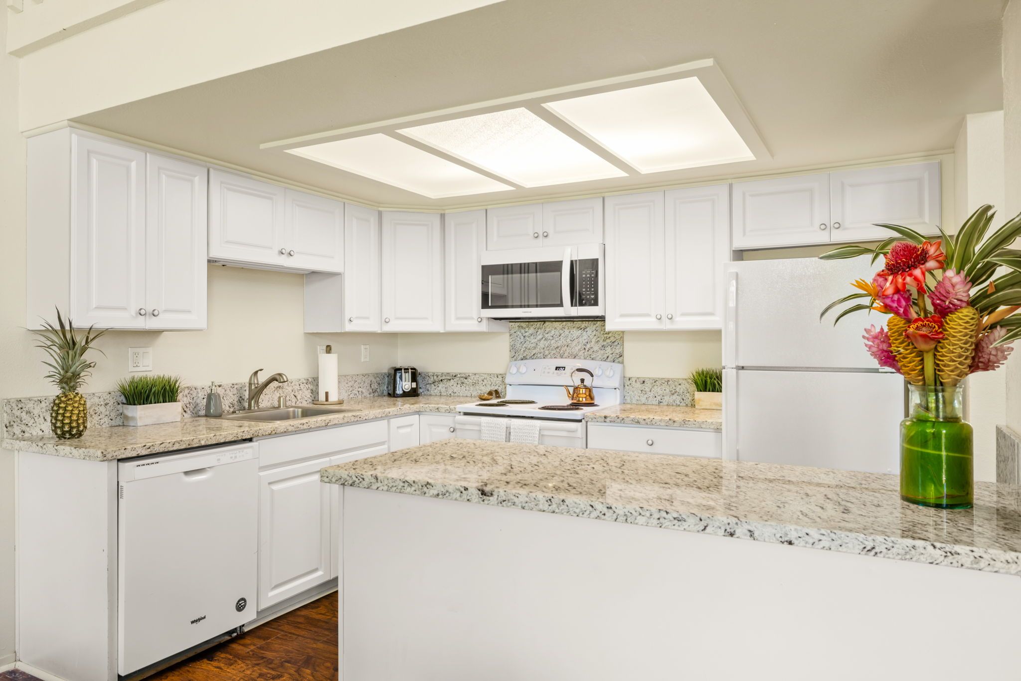 A modern kitchen with white cabinetry, a granite countertop, a stainless steel sink, a microwave, a stove, and a refrigerator.