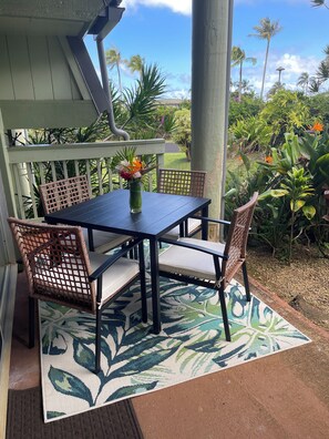 A covered outdoor seating area with a table and chairs, a decorative vase with flowers, and a view of tropical plants and a clear sky. - Hallelujah Condo near Hanalei - 2 Bed 2 Bath (Princeville)