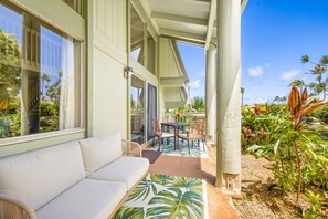 Un espace extérieur aménagé, abrité et doté d’un canapé et de fauteuils, d’une table ornée d’un vase de fleurs et offrant une vue sur une végétation tropicale.