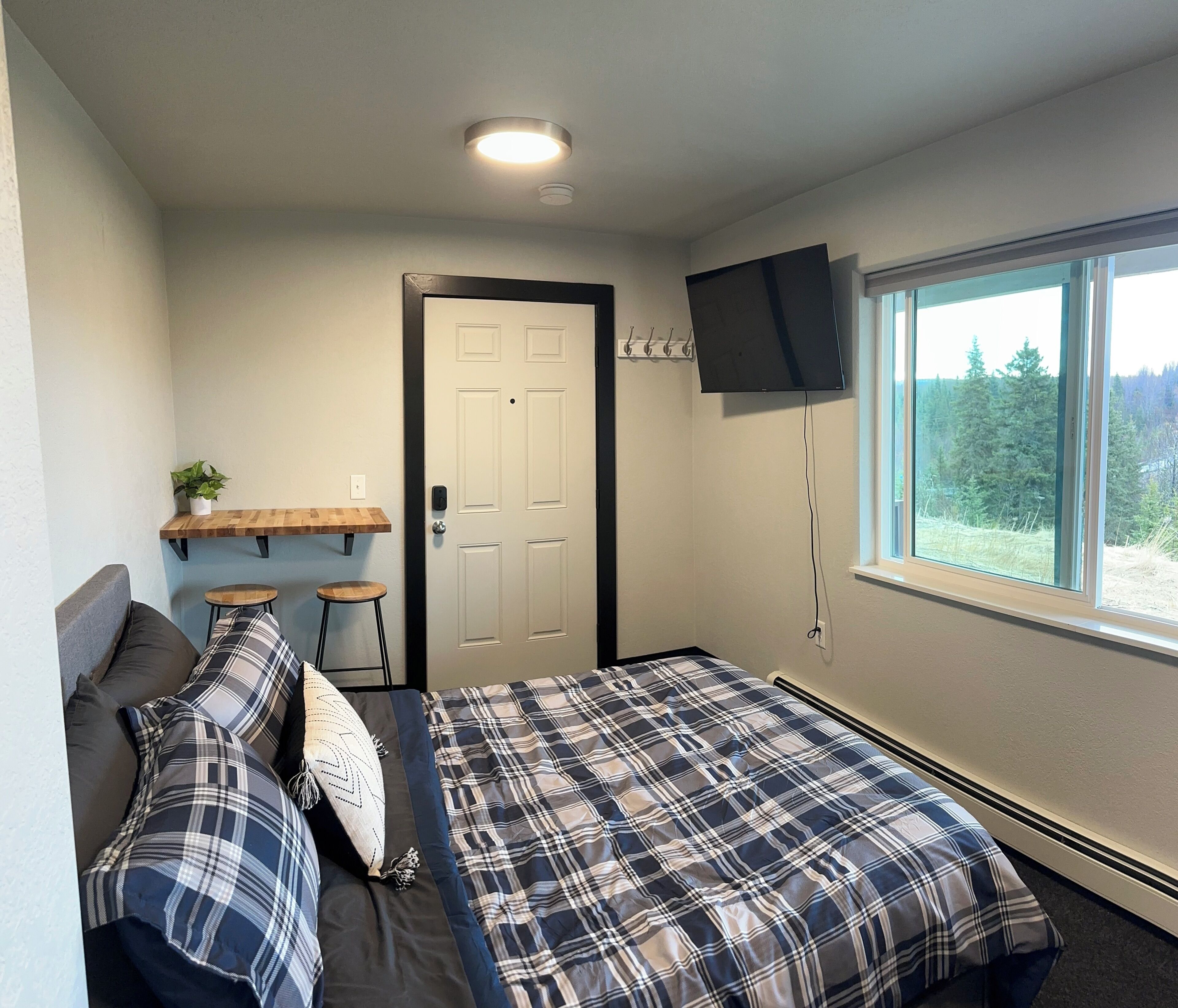 A bedroom with a bed, a TV mounted on the wall, a window with a view of trees, and a door.