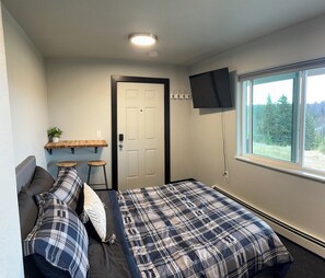 Une chambre à coucher avec un lit, un téléviseur fixé au mur, une fenêtre donnant sur des arbres et une porte.