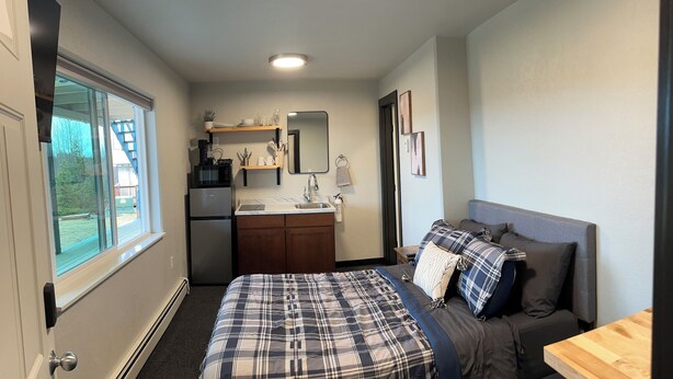 A compact hotel room with a bed, a small desk, a kitchenette, and a window with a view of the outdoors. - ROOM 23. Angry Salmon Inn (Anchor Point)