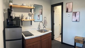 A compact kitchen with a microwave, sink, and wooden cabinets. There is a shelf above the microwave holding dishes and glasses. - ROOM 23. Angry Salmon Inn (Anchor Point)