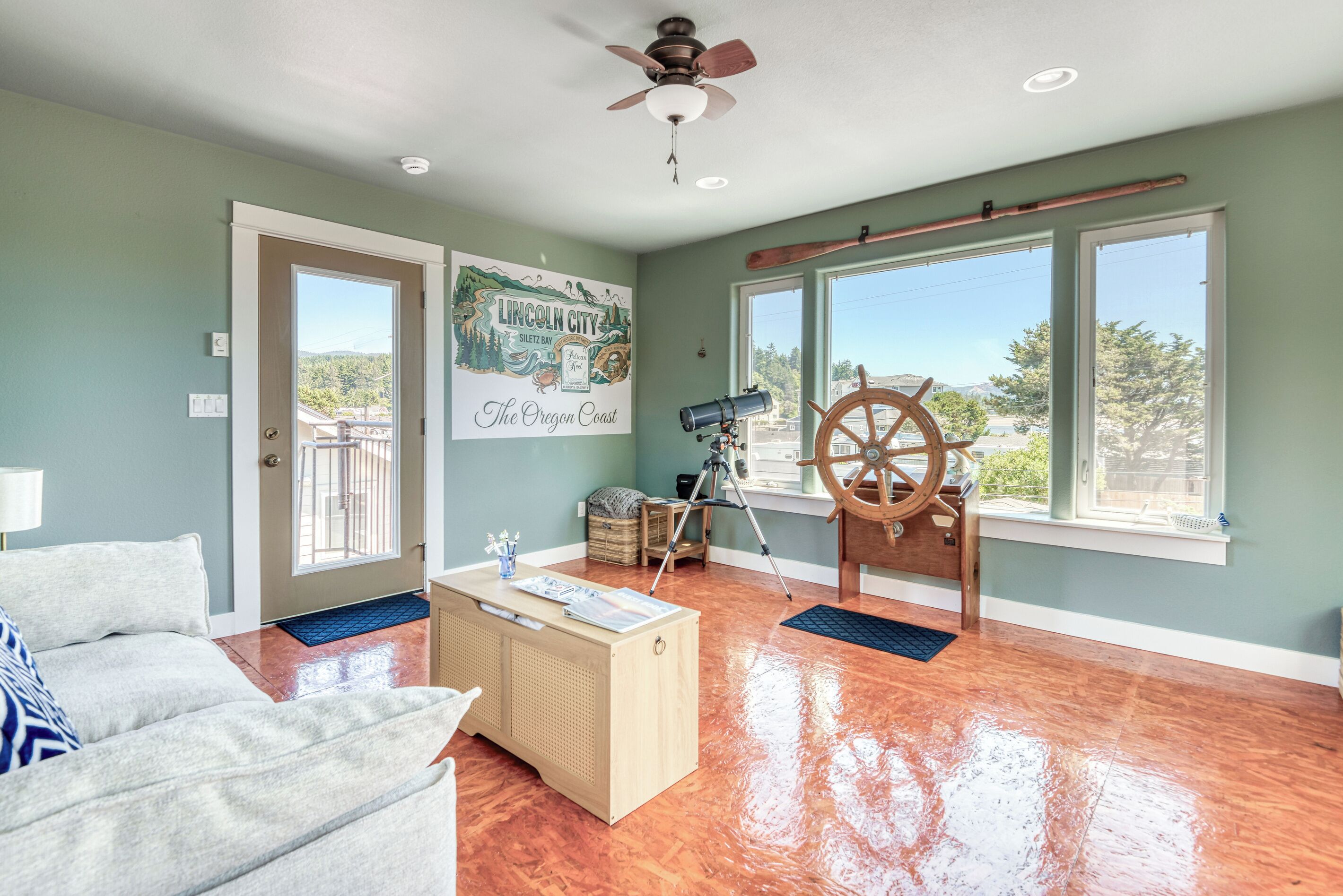 A spacious living room with a wooden floor, a ceiling fan, a large window with a view of the coast, and a sail wheel decoration.