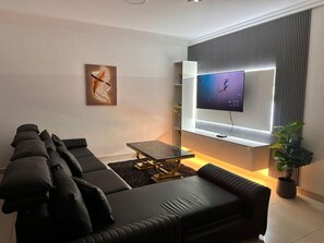 A modern living room with a sectional sofa, a glass coffee table, a flat-screen TV mounted on a wall, and a potted plant. - Charming 3-bedroom apartment with AC in Accra Oyarifa (Accra Oyarifa)