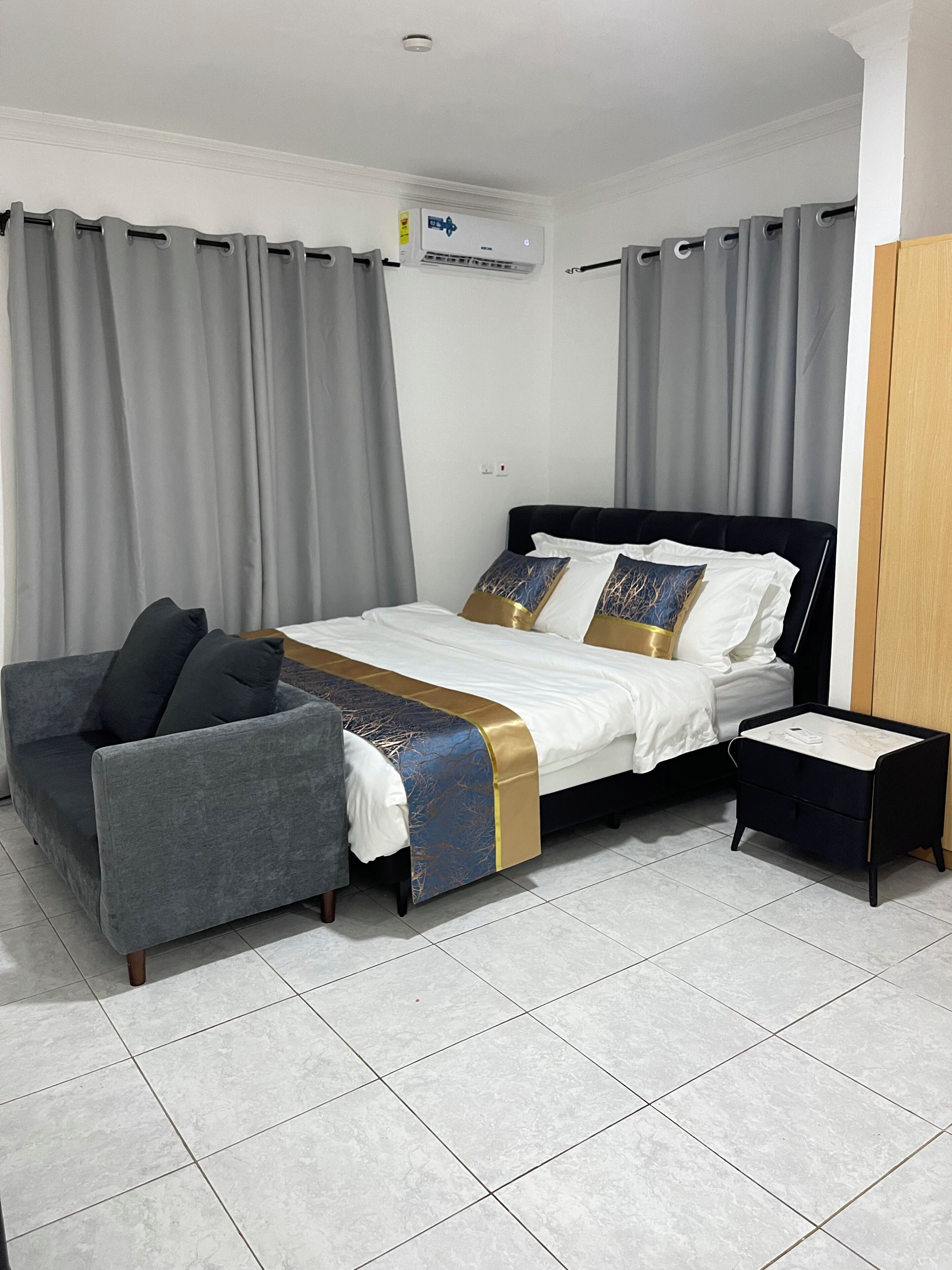 A hotel room with a bed, a sofa, a bedside table, and an air conditioning unit.