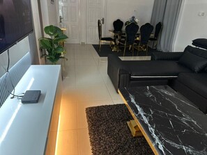 A modern living room with a white TV stand, a black sofa, a marble coffee table, and a dining area with a black table and chairs. - Charming 3-bedroom apartment with AC in Accra Oyarifa (Accra Oyarifa)