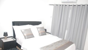 A hotel room with a bed, a nightstand, a lamp, and a window with curtains. - Charming 3-bedroom apartment with AC in Accra Oyarifa (Accra Oyarifa)