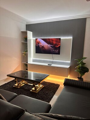 Ein modernes Wohnzimmer mit einem an der Wand befestigten Flachbildfernseher, einem schwarzen Couchtisch mit goldenen Beinen und einem Sofa.