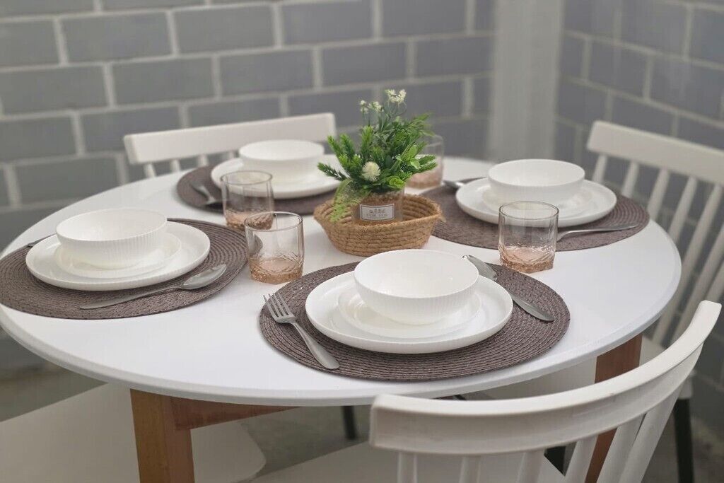 A round dining table with white chairs, set with white plates, cutlery, and glassware, centered with a woven basket containing a small plant.