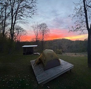 A tent set up on a wooden platform in a grassy area with a small cabin in the background and a forested hill.