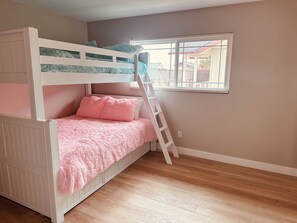 A bunk bed with a pink blanket and fluffy pillows, a wooden ladder, and a window with a view of buildings. - Morro Bay Getaway Beach Retreat! (Morro Bay)