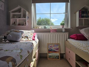 Quarto de criança com beliches, janela com vista e estantes cheias de brinquedos e livros.