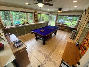 A pool table in a room with large windows, a ceiling fan, and a trash can.