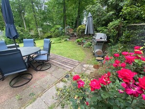 A patio with a table and chairs, a grill, and a garden with roses.