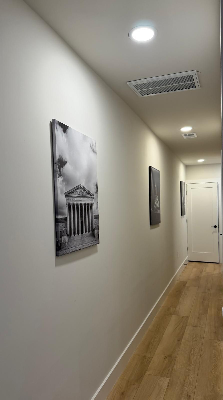 A hallway with a wooden floor, a ceiling vent, and a door at the end.