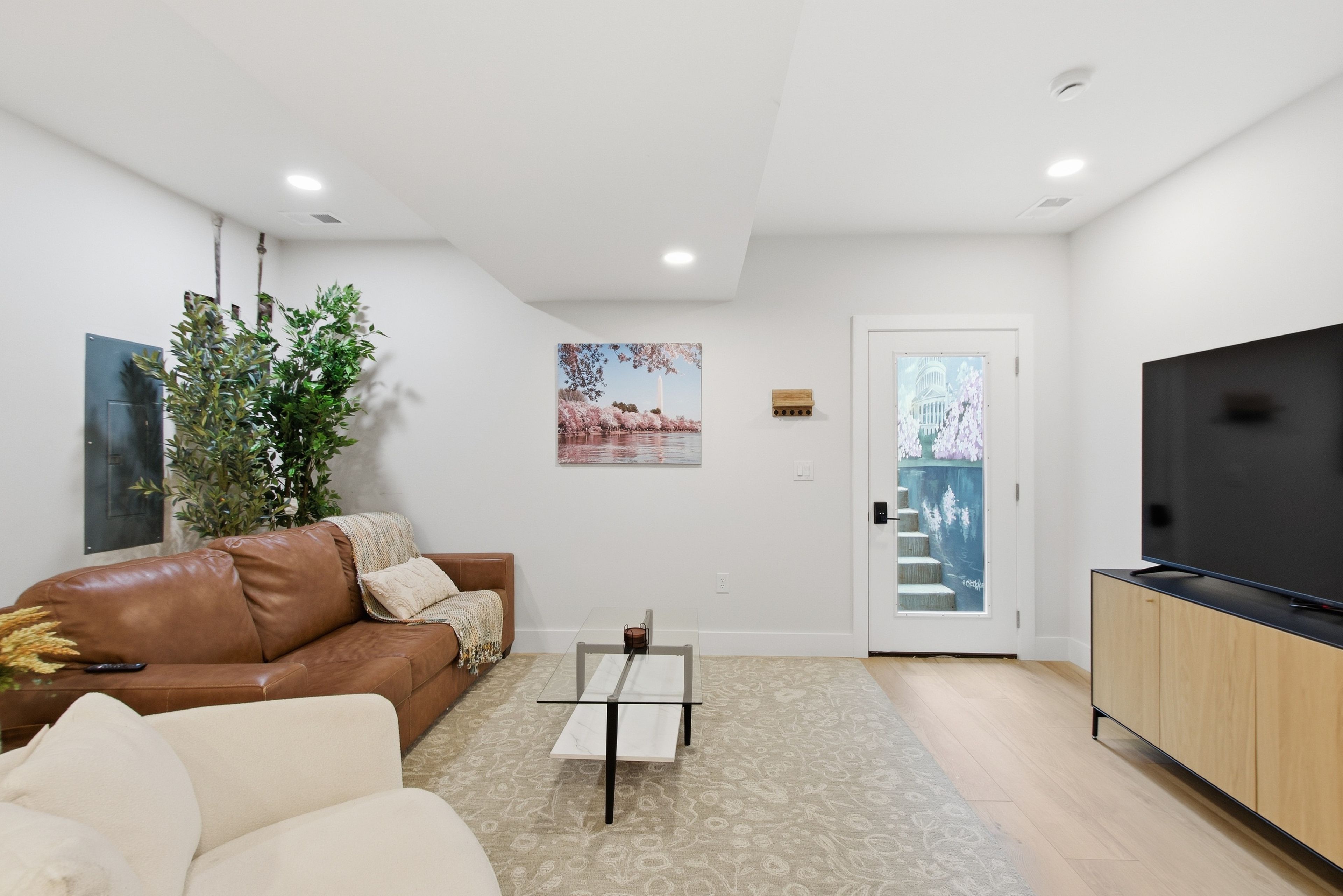 A modern living room with a brown leather sofa, a white armchair, a glass coffee table, a flat-screen TV, and a door with a painting of a building.