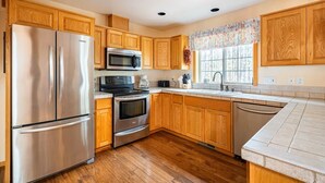 Una cocina con armarios de madera, electrodomésticos de acero inoxidable, una ventana con cortina floral y piso de madera.