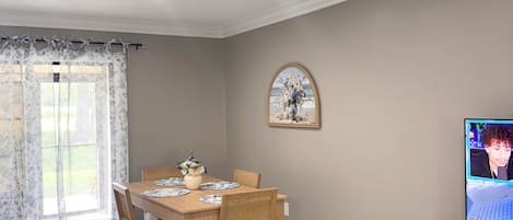 Un comedor con una mesa preparada para cuatro personas, un televisor sobre un mueble bajo y un cuadro en la pared.