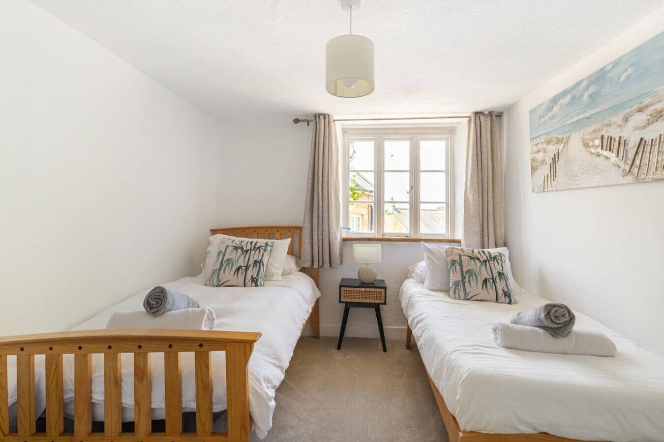 A bedroom with two single beds, a wooden crib, a bedside table with a lamp, a window with curtains, and a beach-themed painting on the wall.