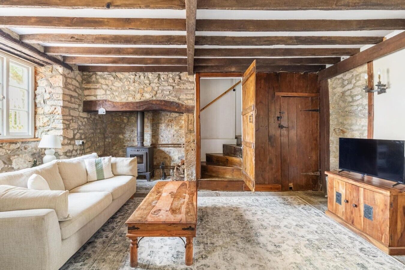 Un salon rustique avec un mur en pierre, un canapé en bois, une table en bois, un téléviseur posé sur un meuble en bois et un escalier menant à l’étage supérieur.