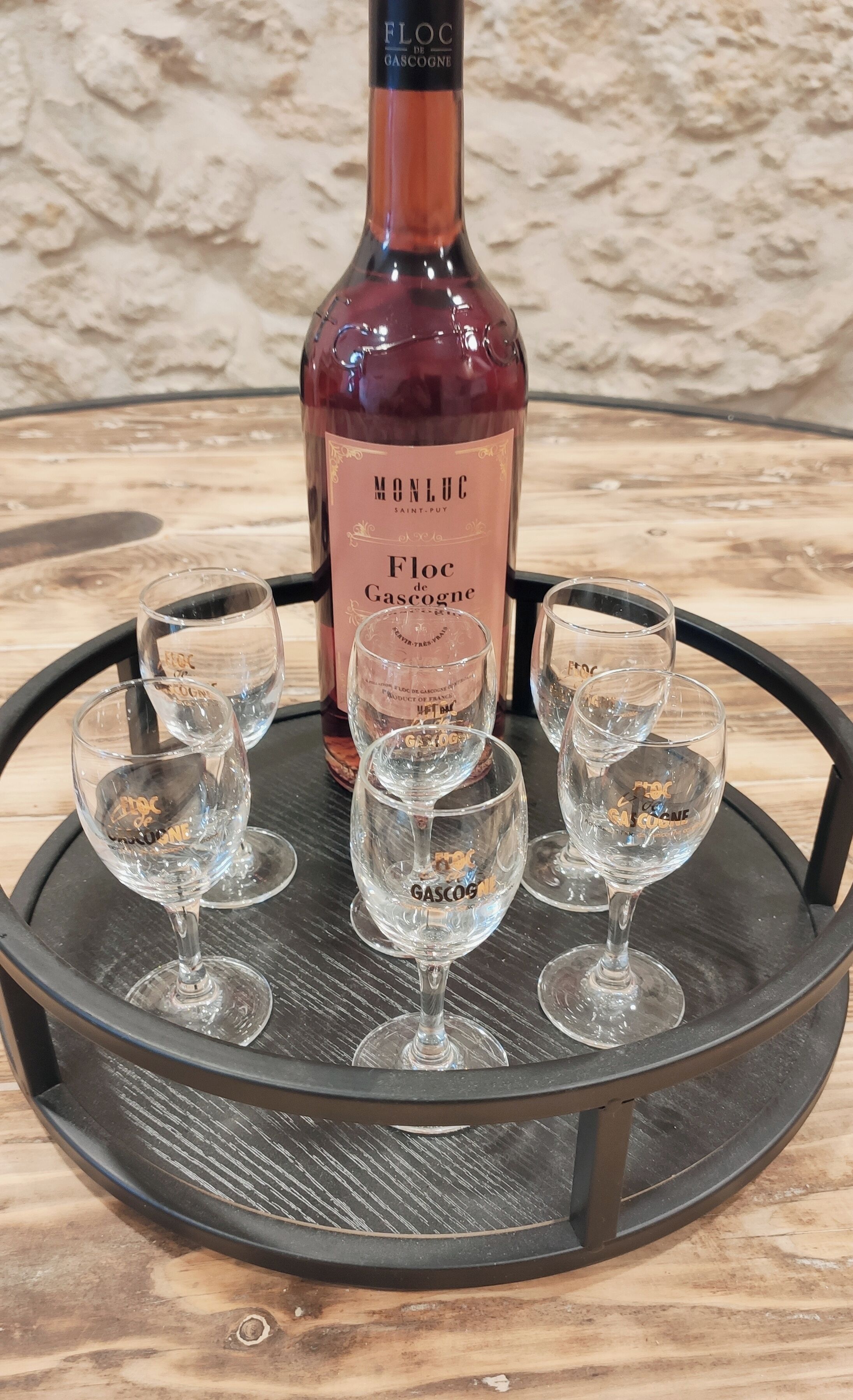 Une bouteille de vin Monluc Floc de Gascogne, accompagnée de cinq verres, est posée sur un plateau rond noir.
