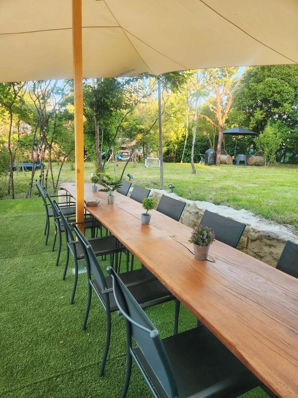 Un área de comedor al aire libre con una larga mesa de madera y sillas, rodeada de follaje y árboles.