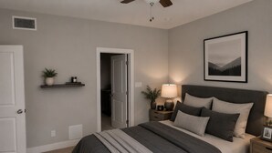 Quarto com cama, mesas de cabeceira, abajur, quadro pendurado na parede e porta que dá para outro cômodo.