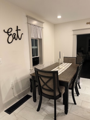 A dining room with a wooden table, chairs, a window with white curtains, a wall-mounted sign, and a vent.