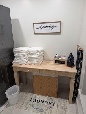 Un coin buanderie avec une pile de serviettes blanches, une citation inspirante encadrée, un panier à linge et un aspirateur.