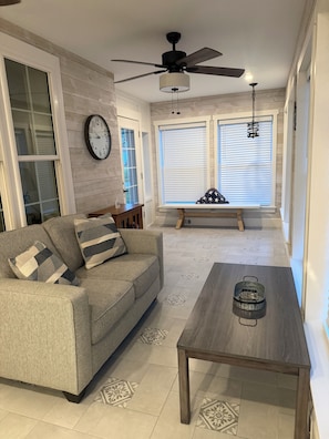 Un salon comprenant un canapé, une table basse, un ventilateur de plafond, une horloge et une fenêtre avec des stores.