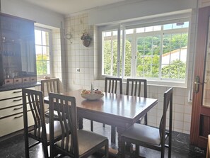 A dining area with a wooden table and chairs, a kitchen with white tiles, and a window with a view of greenery. - La Coqueta Apartment 2034 (San Miguel de Meruelo)