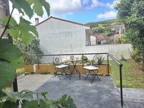 Een terras met twee metalen stoelen en een kleine tafel, omgeven door een lage gele muur en begroeiing.