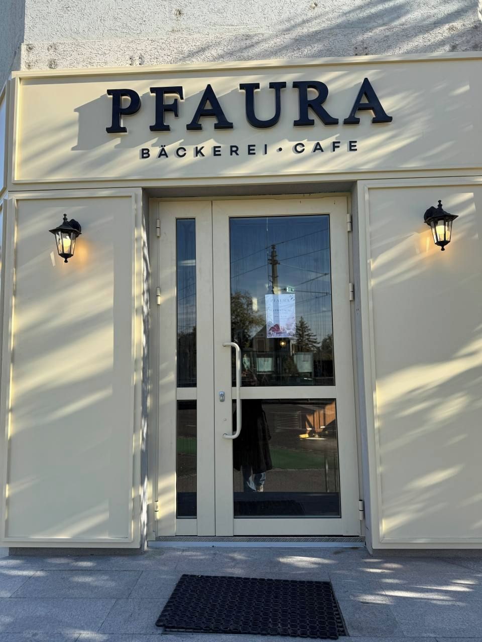 La entrada a PFAURA BÄCKEREI CAFE con un escaparate de vidrio y dos lámparas fijadas a la pared.